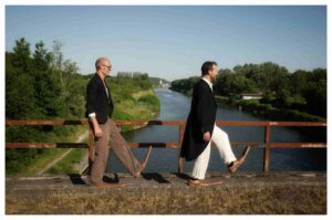 Les comédiens marchent sur un pont au-dessus d'une rivière. Ils portent des grandes chaussures.