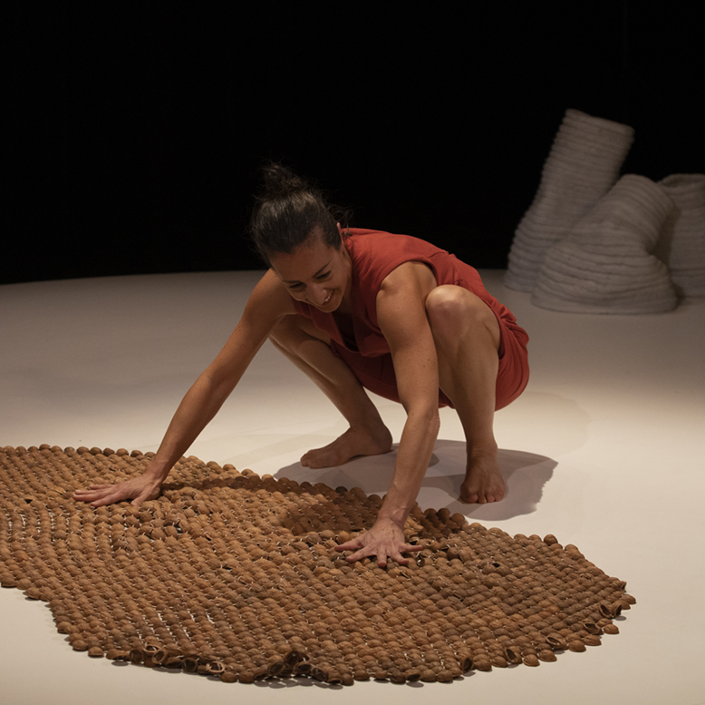 La comédienne est accroupie au sol et touche un tapis de coquilles de noix.